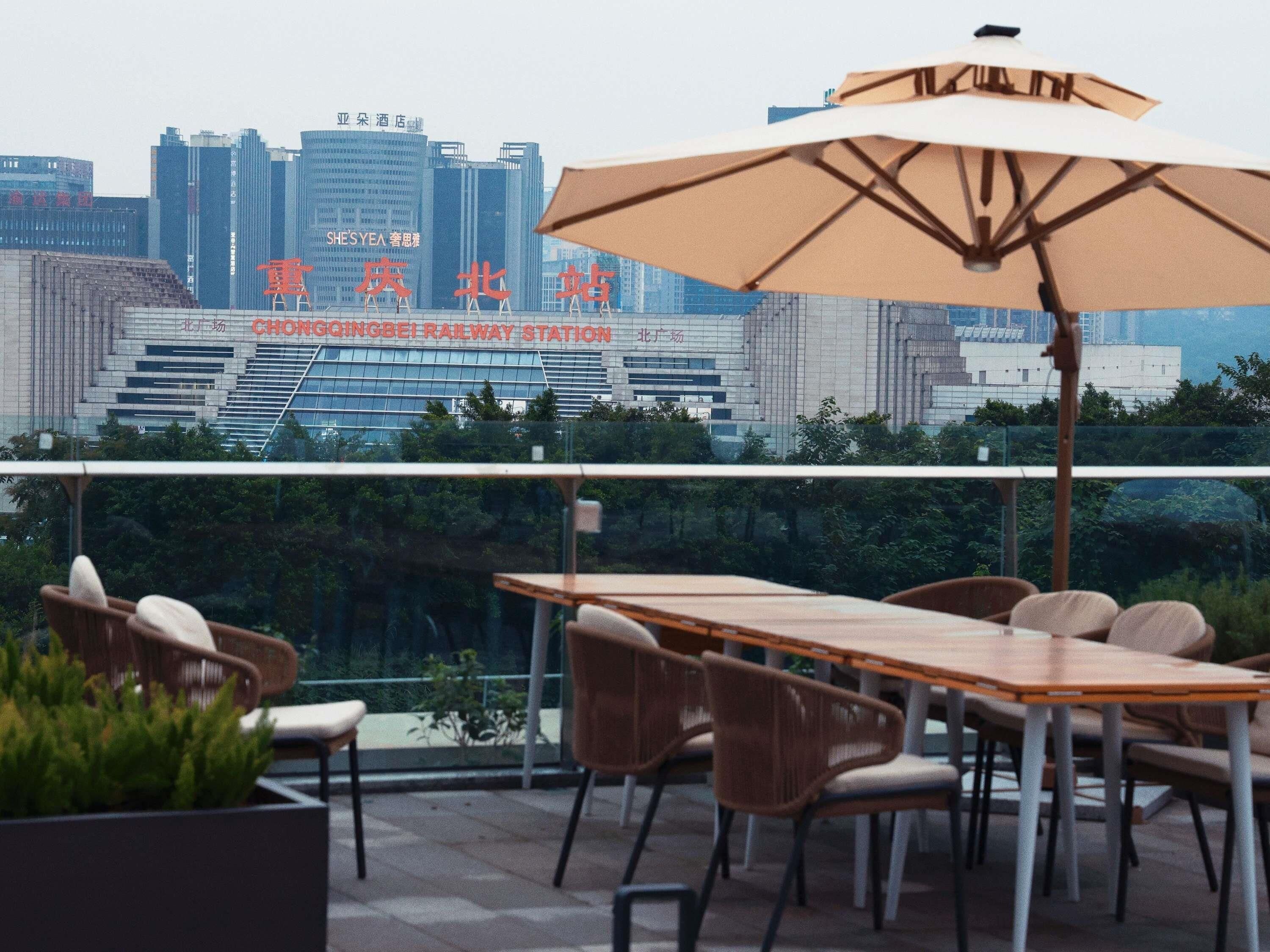 A rooftop terrace with a table and chairs, a large umbrella, and a view of the Chongqing Railway Station in the background.