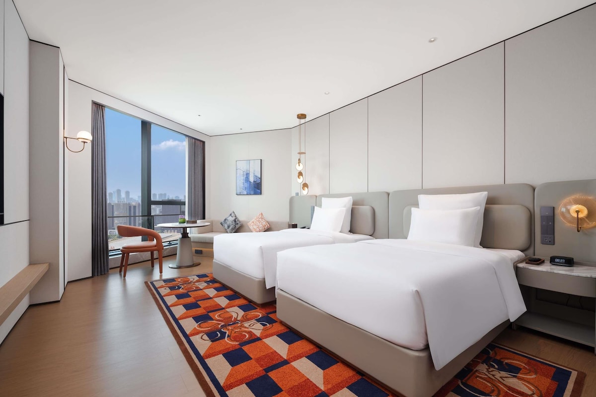 Ein modernes Hotelzimmer mit zwei Betten, einem Schreibtisch, einem Stuhl, einem großen Fenster mit Blick auf die Stadt und einem gemusterten Teppich.