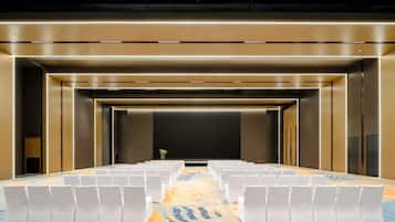 Un salón de conferencias con filas de sillas blancas, piso alfombrado y una pantalla oscura al frente.