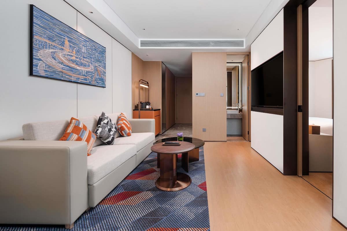 Ein modernes Hotelzimmer mit einer Couch, einem Couchtisch, einem GemĂ€lde, einem Spiegel, einem Fernseher und einem sichtbaren Badezimmer hinter einer offenen TĂŒr.