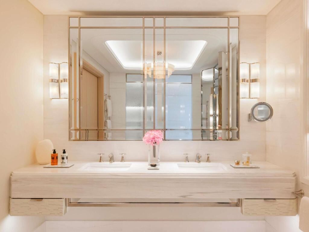 A bathroom with a large mirror, two sinks, a vase with flowers, and wall sconces.