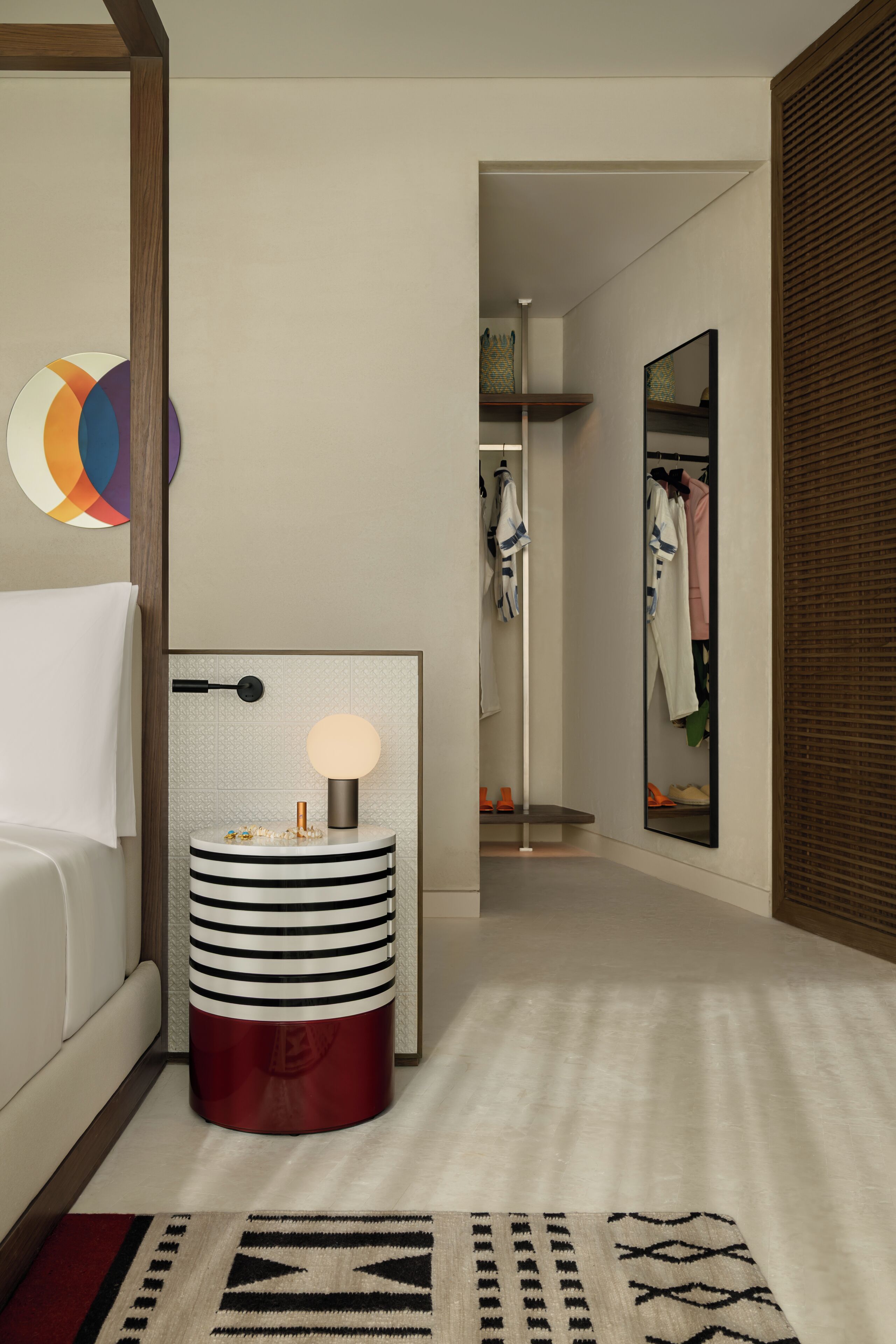 Moderni hotellihuone, jossa on sänky, yöpöytä lampun kanssa, peili ja vaatekaappi.