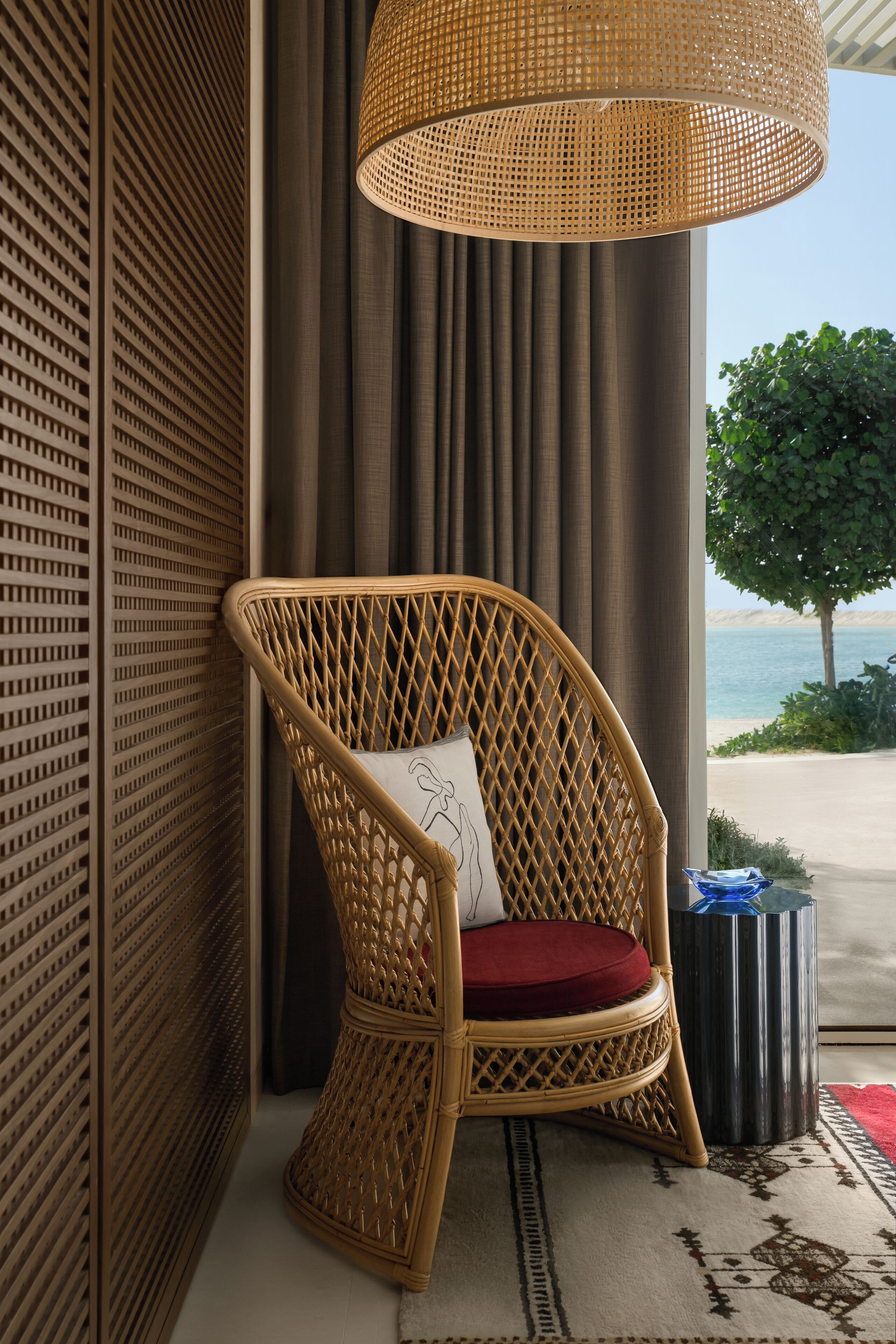 A wicker chair with a red cushion, a woven lamp, and a view of the sea.