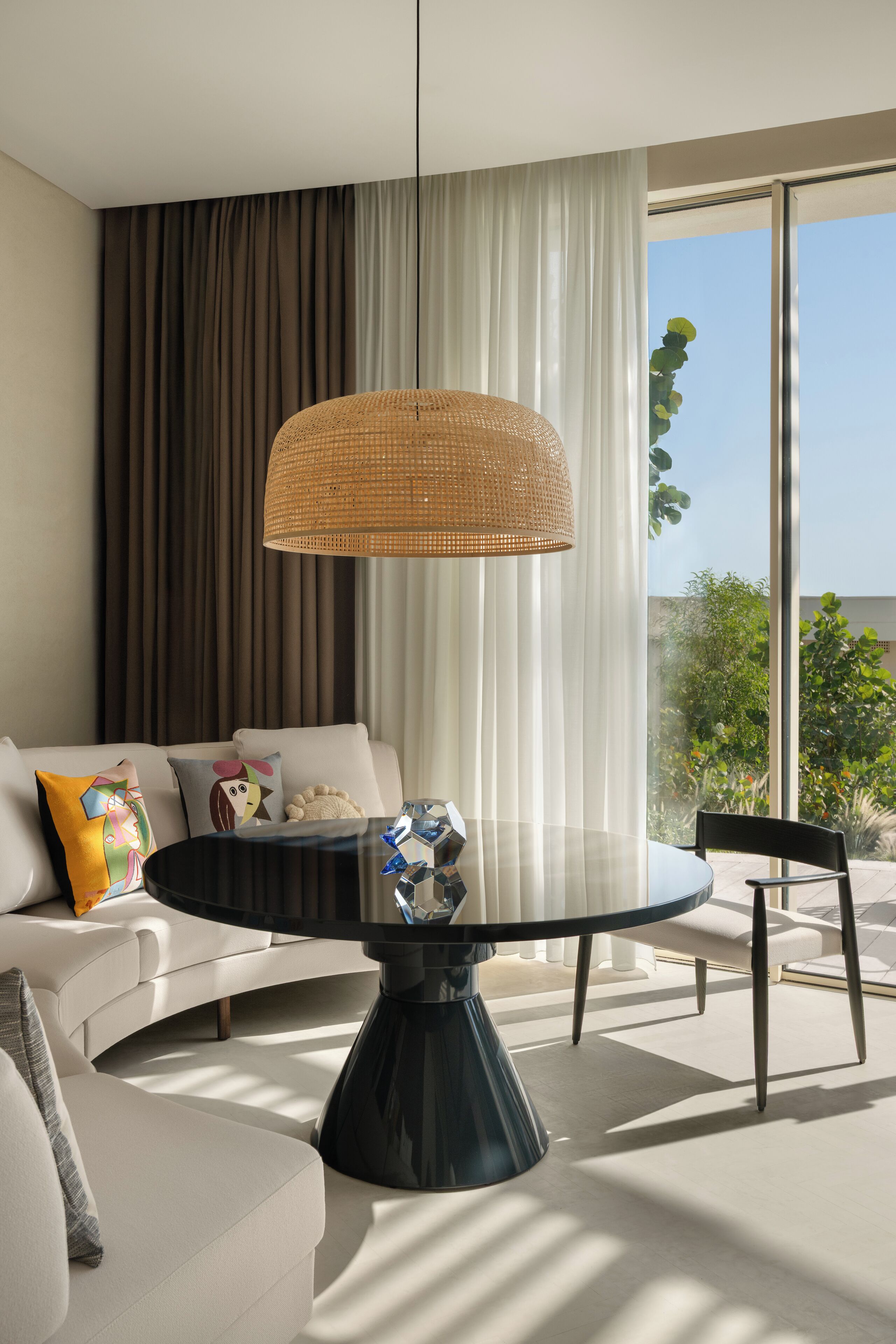 A modern living room with a round glass coffee table, a woven pendant light, a white sofa with colorful cushions, and a view of greenery through large windows.