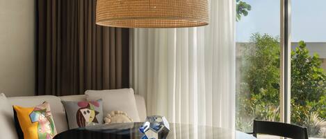 A modern living room with a round glass coffee table, a woven pendant light, a white sofa with colorful cushions, and a view of greenery through large windows.