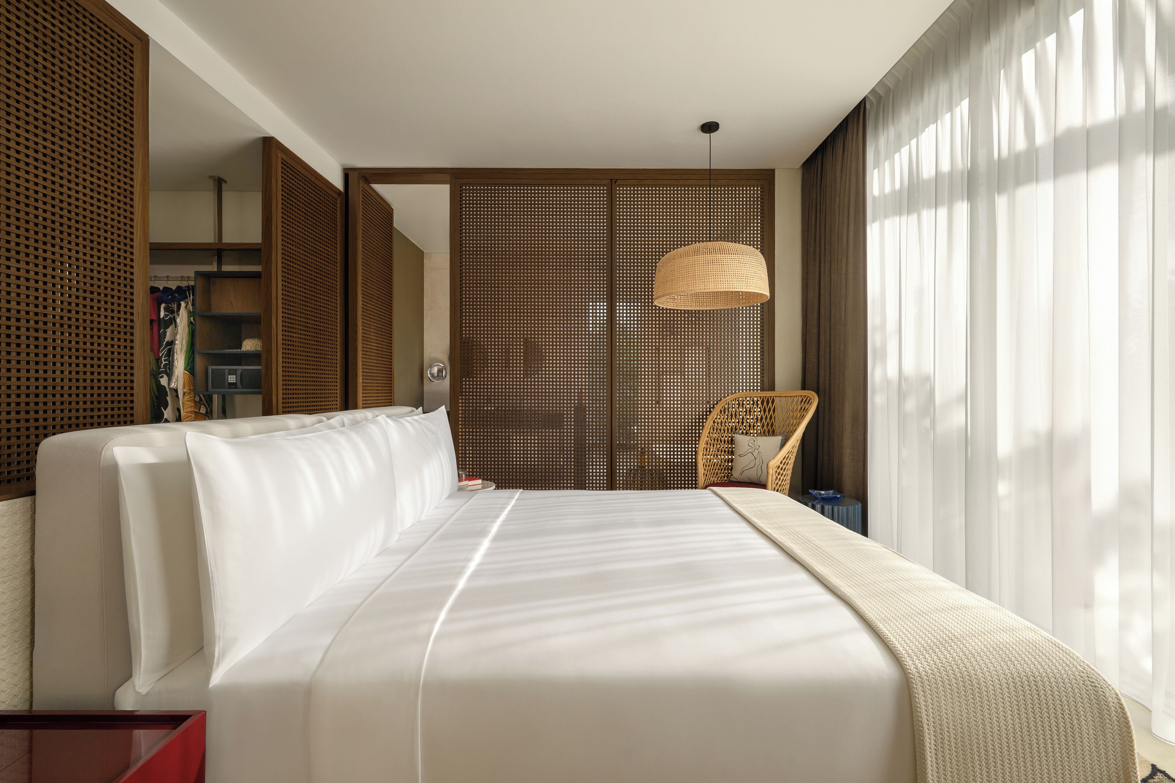 A hotel room with a large bed, a wicker chair, a wardrobe, and a large window with sheer curtains.
