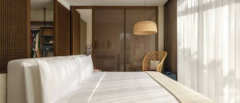 A hotel room with a large bed, a wicker chair, a wardrobe, and a large window with sheer curtains.