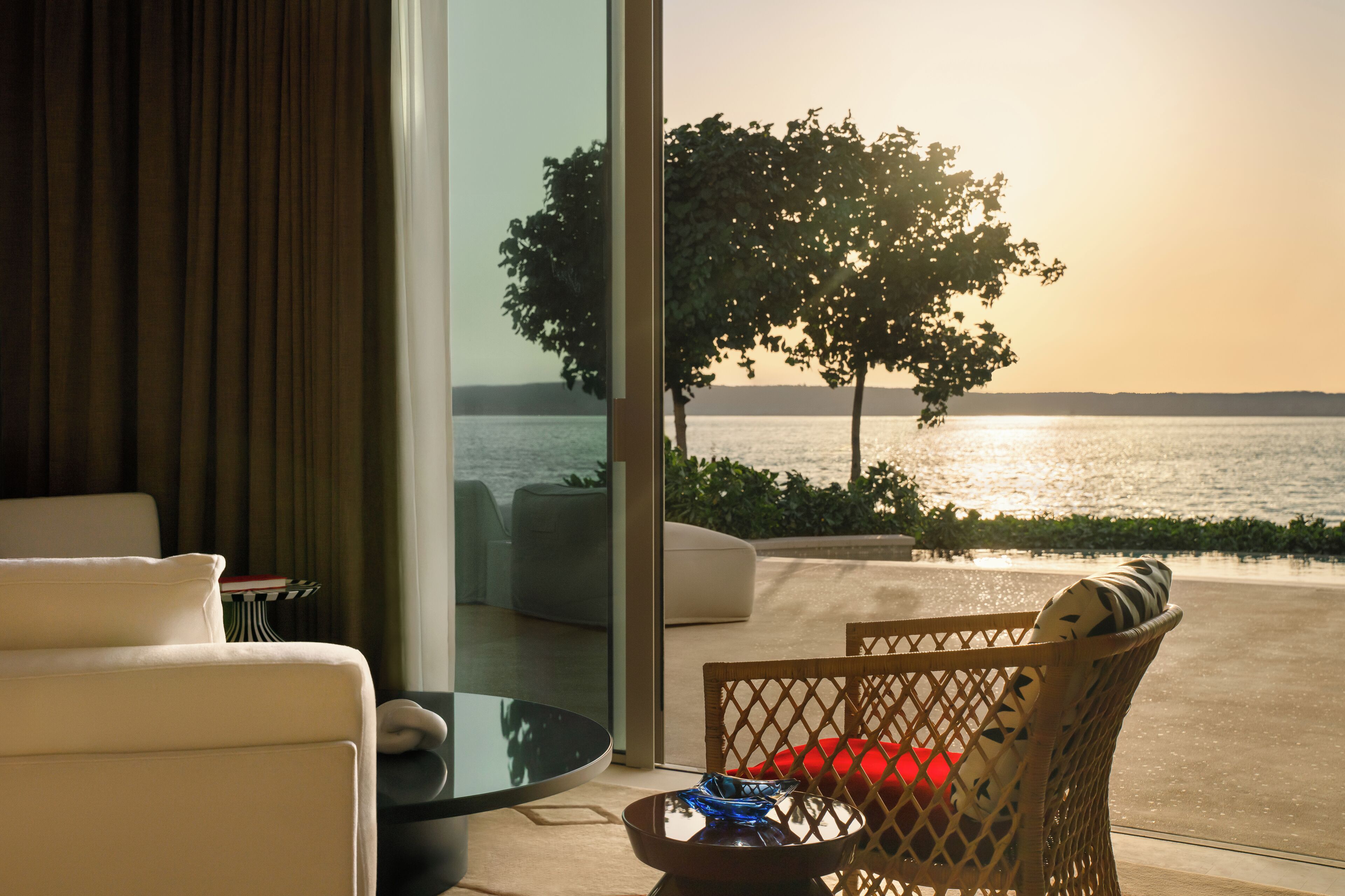 A modern living room with a glass door leading to a balcony overlooking a body of water and a tree.