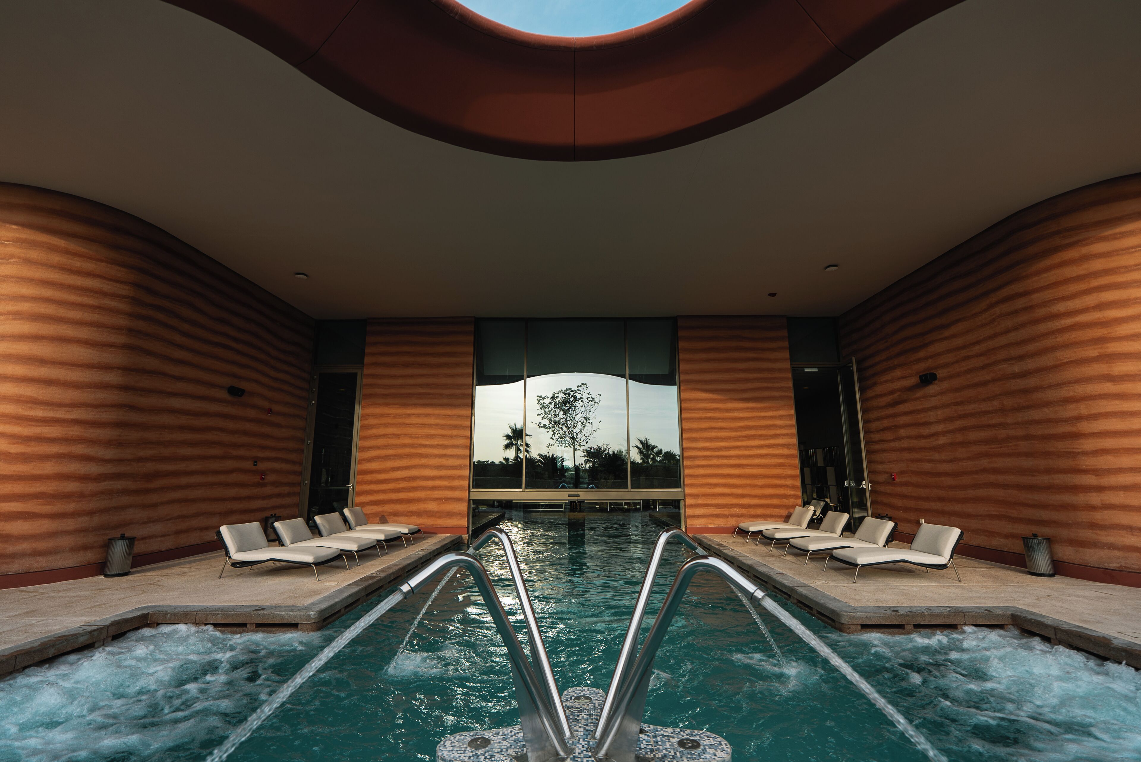 Piscina interior moderna con sillones de descanso, vista a través de un amplio ventanal y un diseño de techo original.