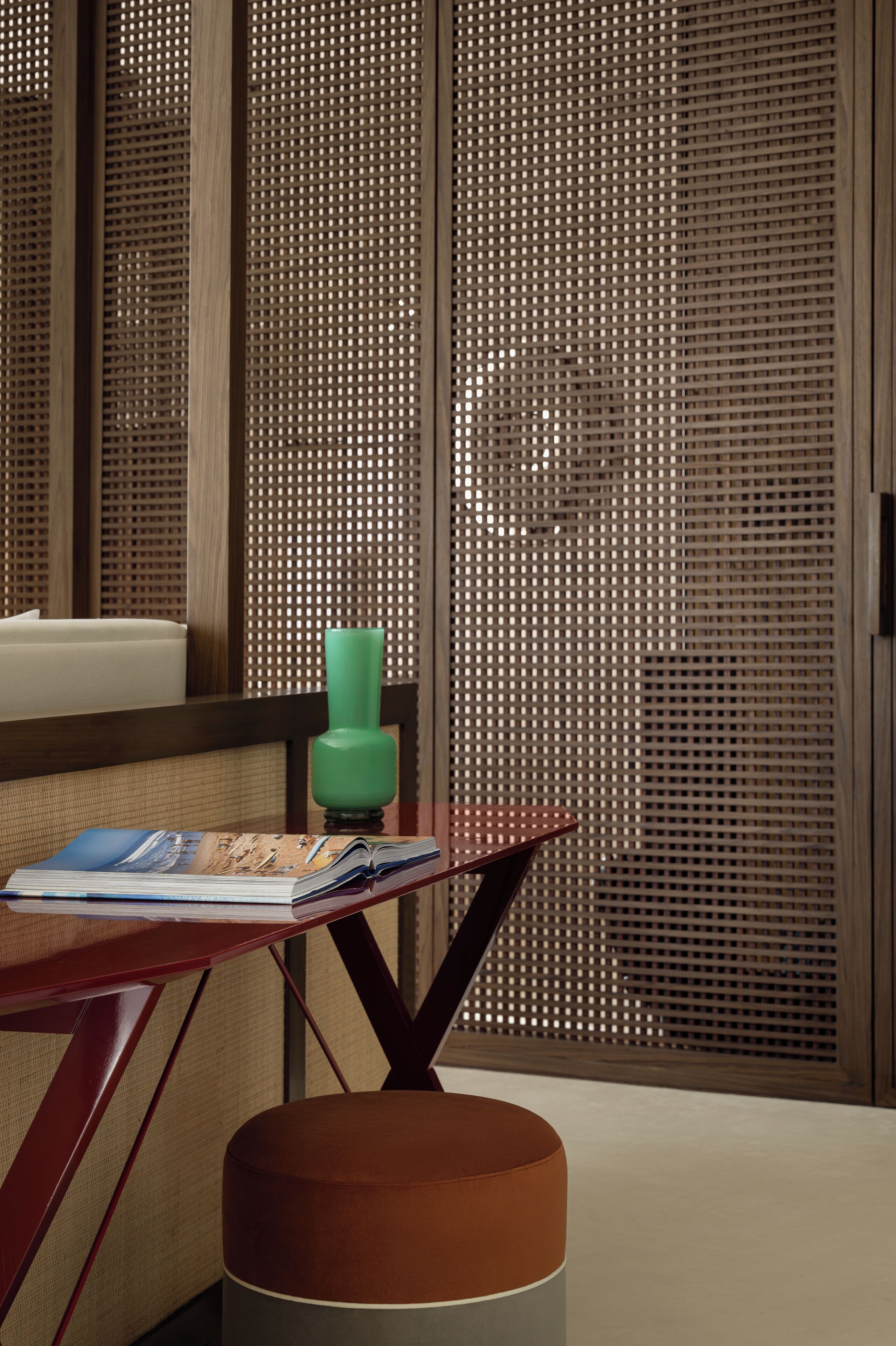 A room with a wooden partition, a small table with a book, a green vase, and a brown ottoman.