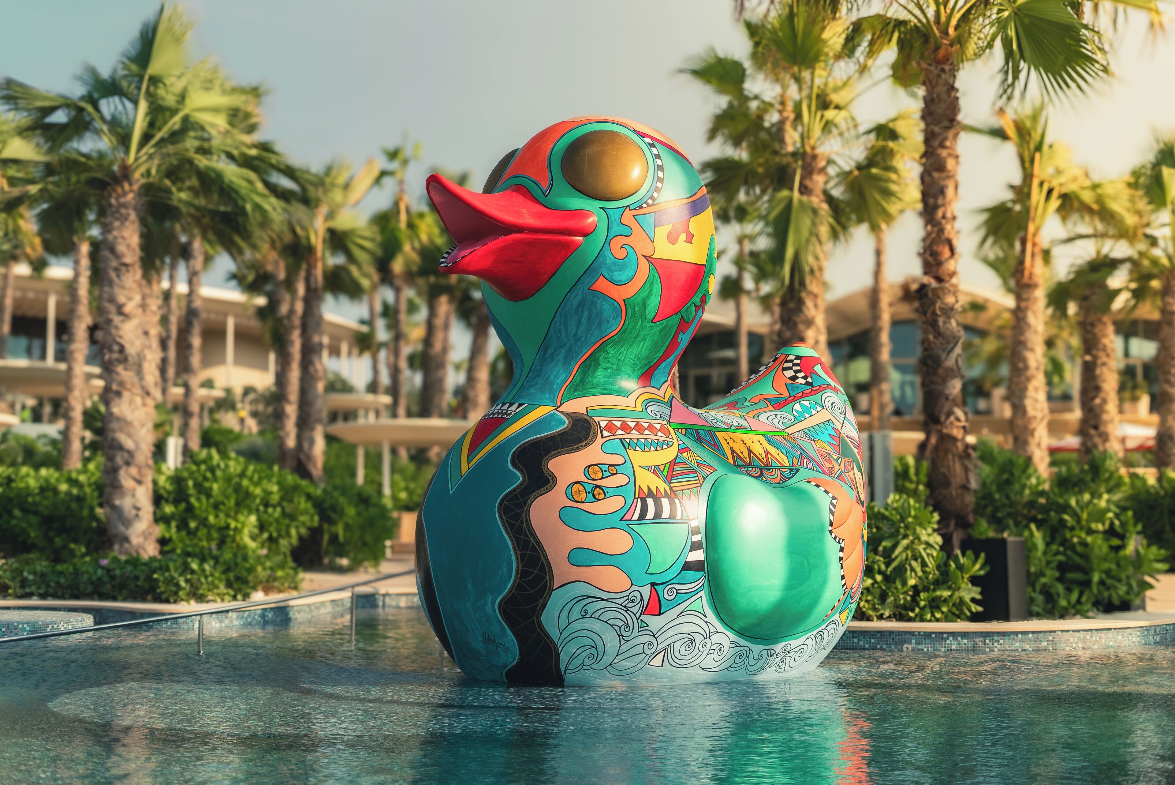 A colorful sculpture in a pool surrounded by palm trees and a building in the background.