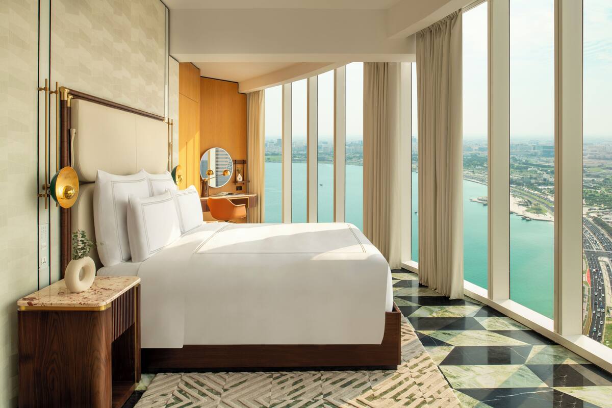 Una habitación de hotel moderna con una cama grande, una mesita de noche, un espejo y vistas a un paisaje urbano y al agua.