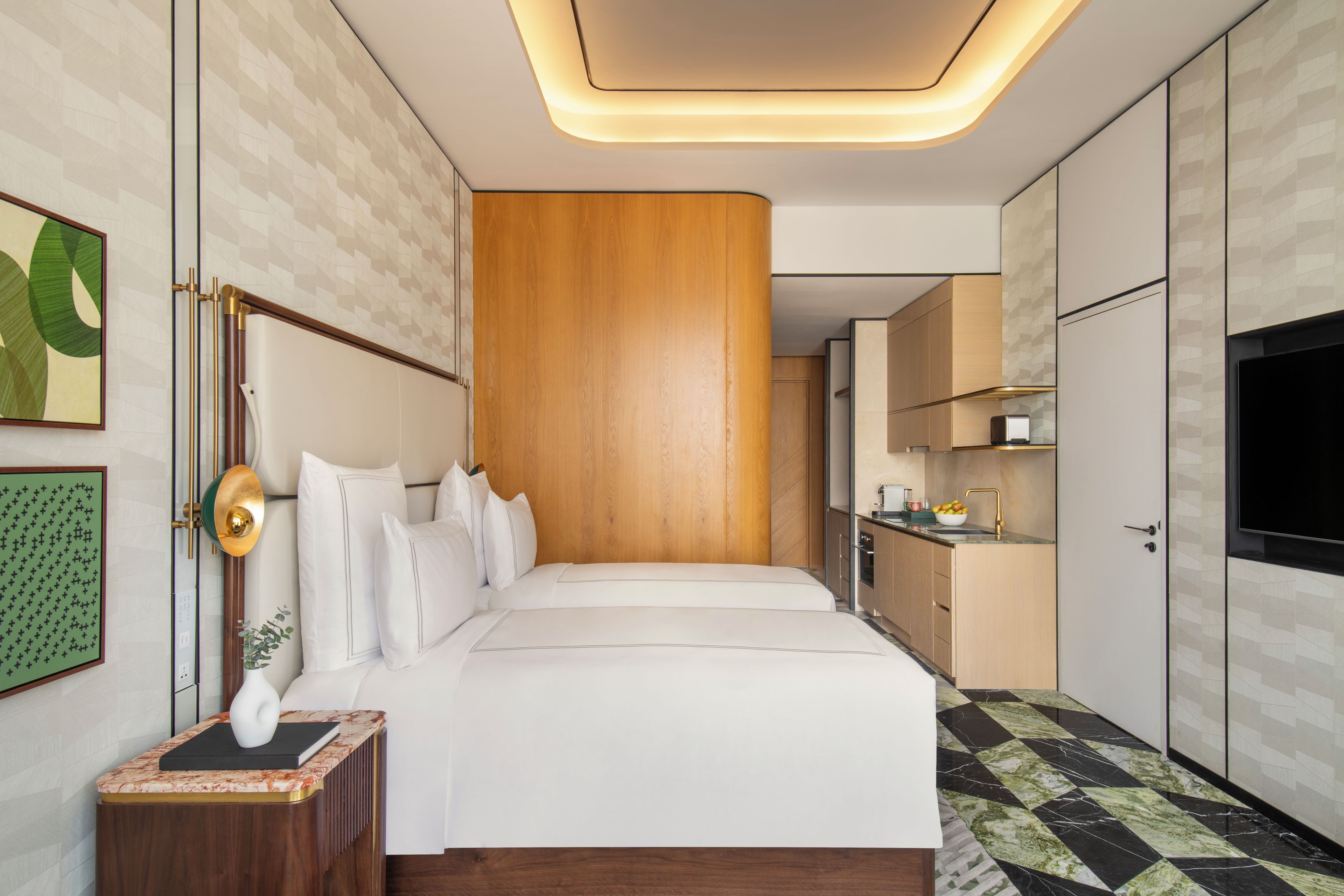 A modern hotel room with a large bed, a bedside table with a lamp, a TV, and a kitchen area with cabinets and a countertop.