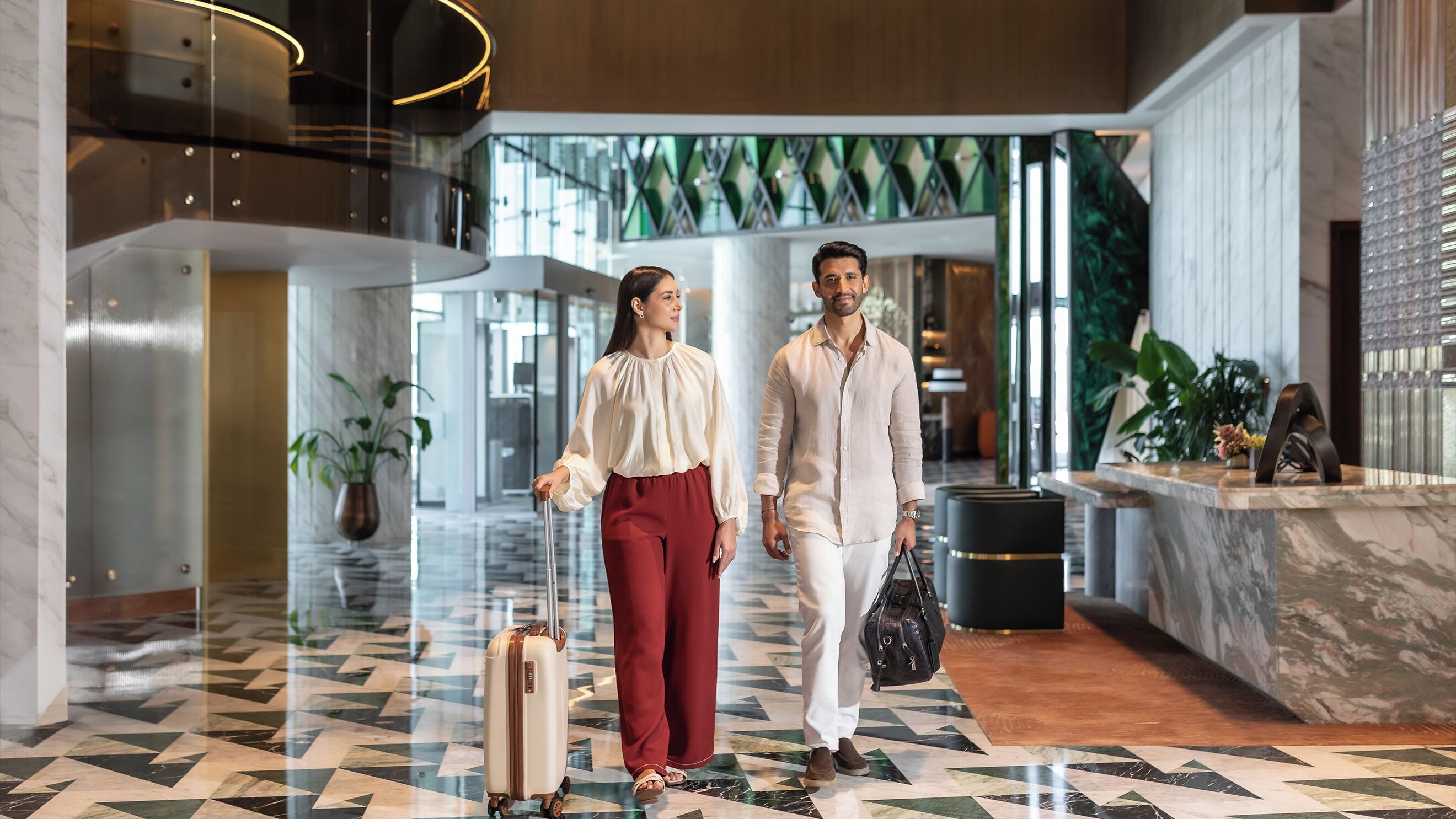 A modern lobby with a geometric tile floor, a glass elevator, and a reception desk with a plant.