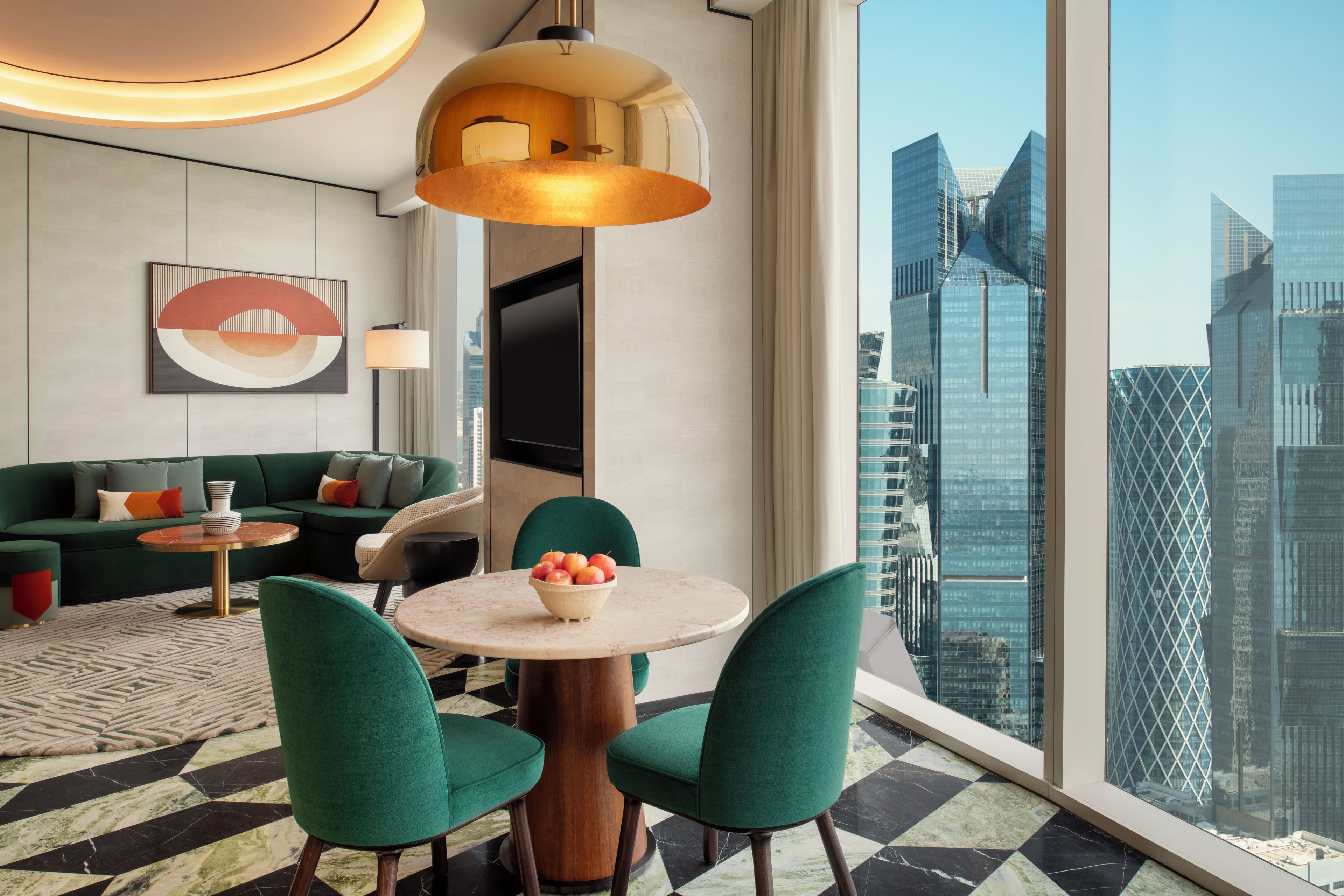 A modern hotel room with a large city view, a round dining table, green upholstered chairs, a sofa with cushions, and a contemporary light fixture.