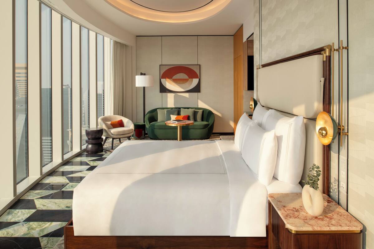Una habitación de hotel moderna con una cama grande, un sillón verde, una mesita y vistas a la ciudad a través de amplios ventanales.