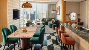 Une chambre d’hôtel moderne dotée d’un coin repas, d’un ensemble de canapés, d’un comptoir de cuisine et d’une grande fenêtre donnant sur la ville.