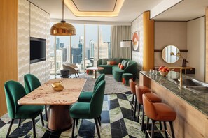 Una habitación de hotel moderna con zona de comedor, un conjunto de sofás, una encimera de cocina y un amplio ventanal con vistas a la ciudad.