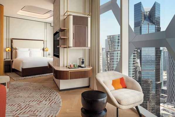 Une chambre d’hôtel moderne dotée d’un grand lit, d’un coin salon avec un fauteuil et une petite table, et offrant une vue sur le paysage urbain grâce à de grandes fenêtres.