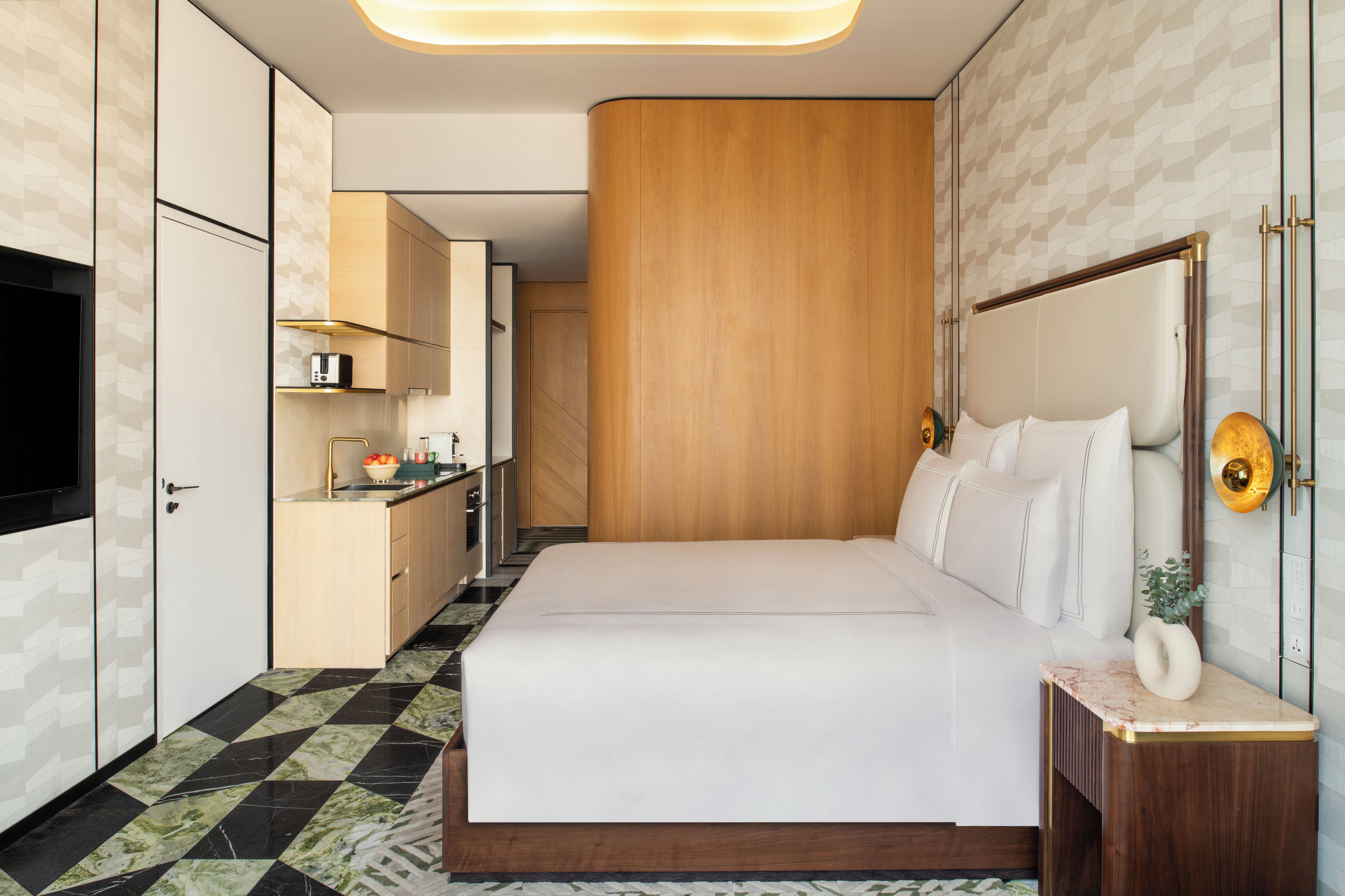 Une chambre d’hôtel moderne avec un grand lit, une tête de lit en bois, une petite table de chevet et un téléviseur fixé au mur.