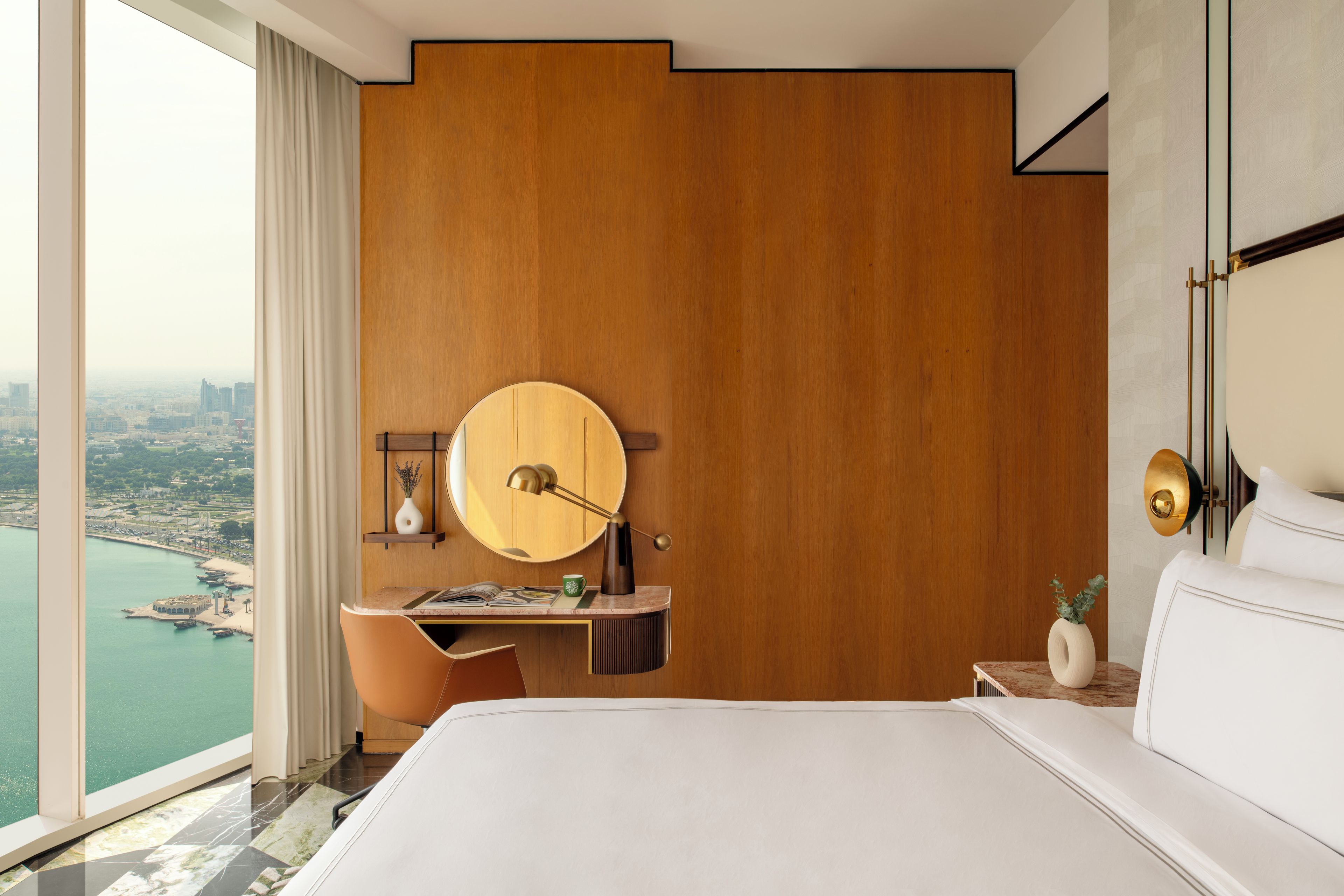Une chambre avec un grand lit, un bureau avec une chaise, un miroir rond et une vue sur le paysage urbain.