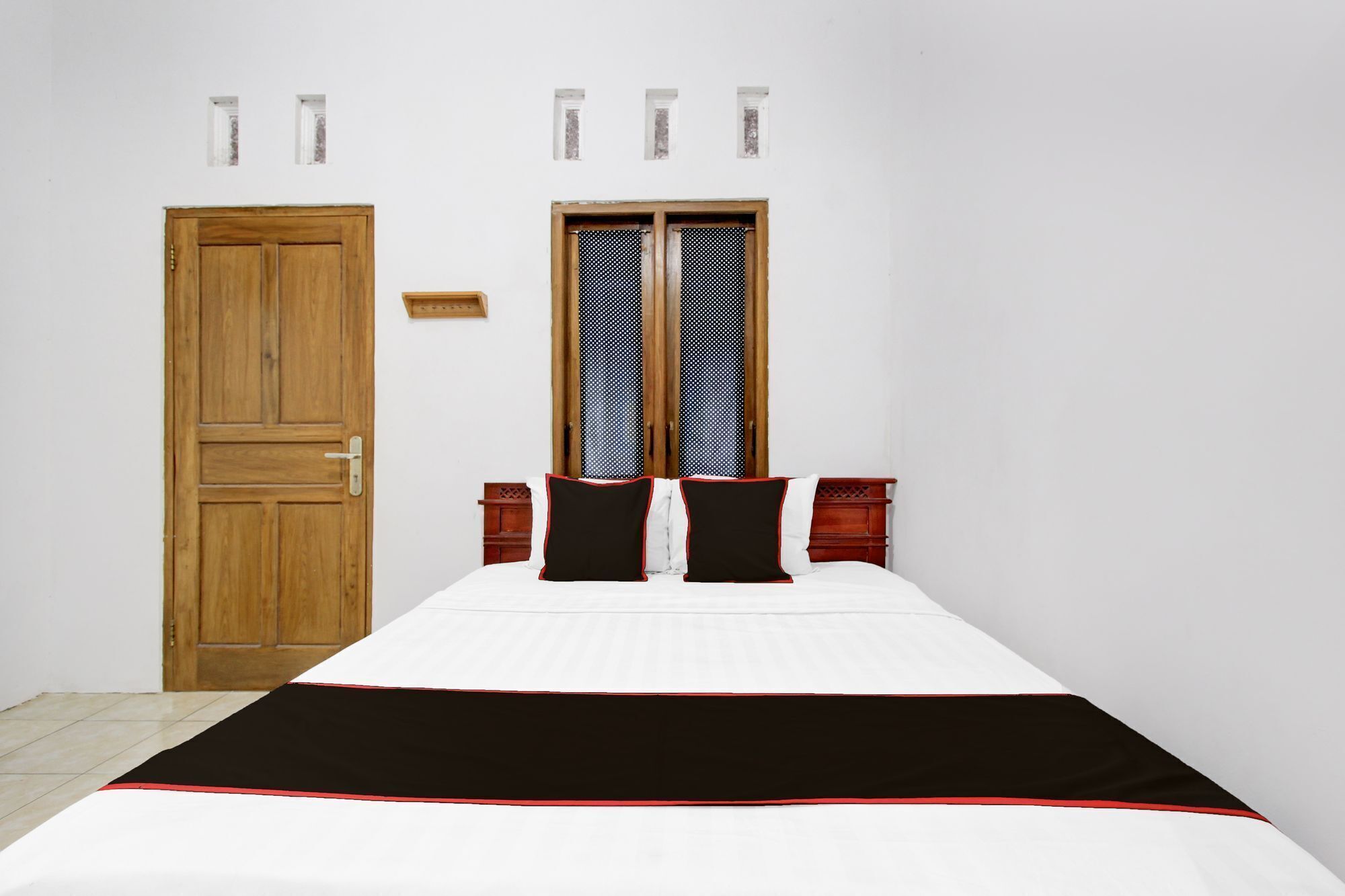 A bedroom with a wooden door, a bed with white bedding and black and red pillows, and a window with a patterned curtain.