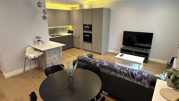 Ein modernes Wohnzimmer mit Esstisch, schwarzen Stühlen, einer Couch, einem Fernseher und einer Küche im Hintergrund.