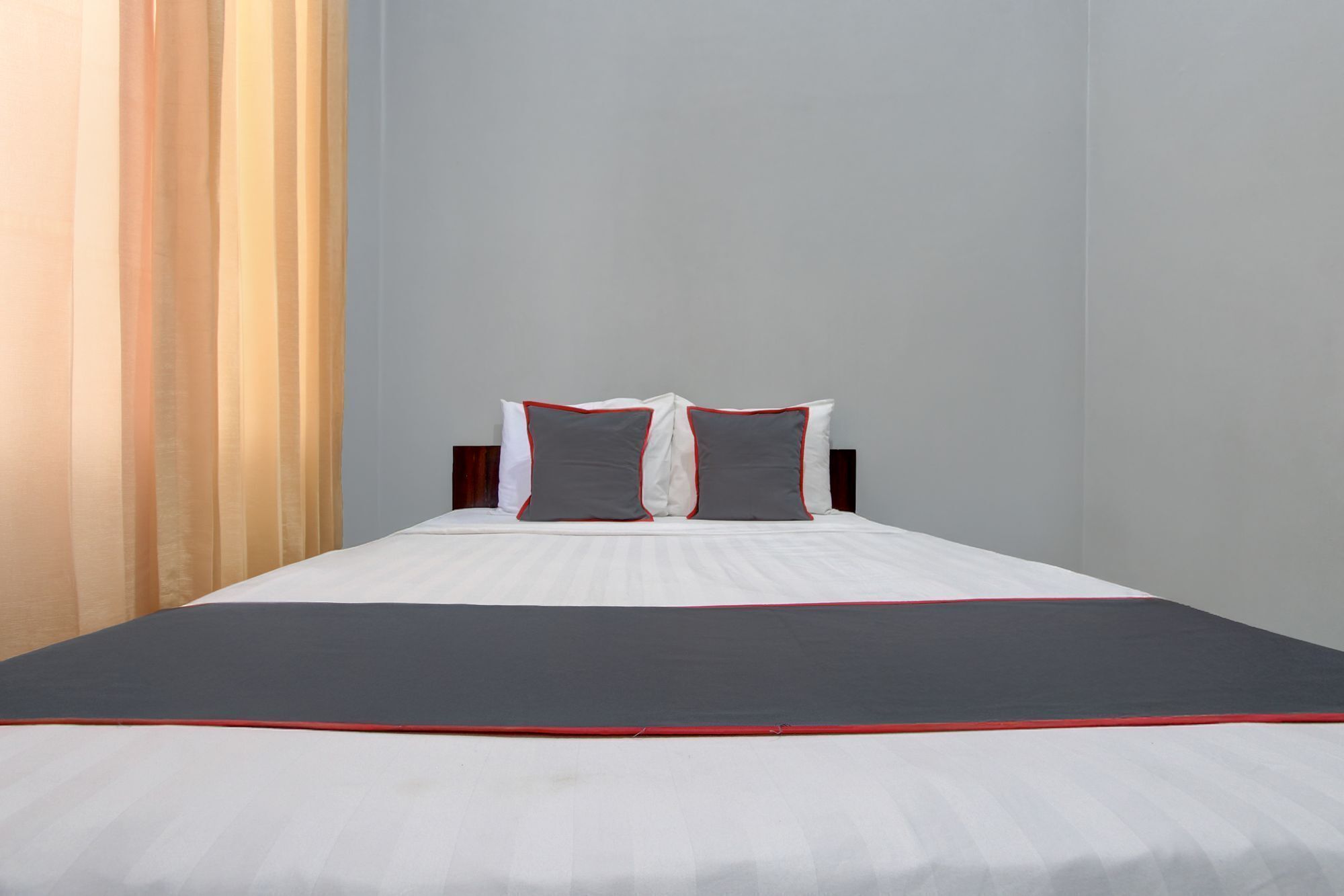 Ein ordentlich bezogenes Bett mit weißer und grauer Bettwäsche, ein dunkelroter Akzent am Fußende und ein Holz-Kopfteil.