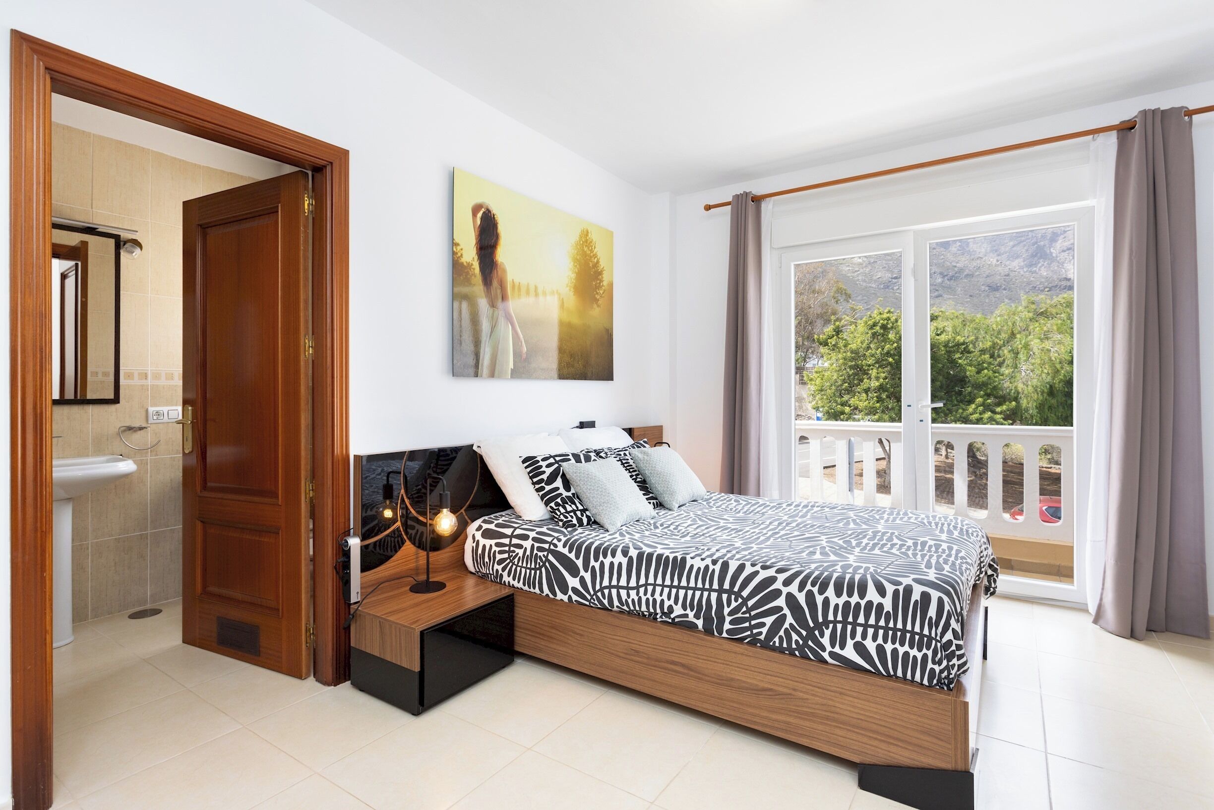 A bedroom with a bed, a nightstand, a wall-mounted lamp, a painting, a window with curtains, and a view of greenery outside.