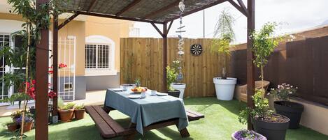 Un patio techado con una mesa lista para dos personas, rodeado de plantas en macetas y una valla de madera.