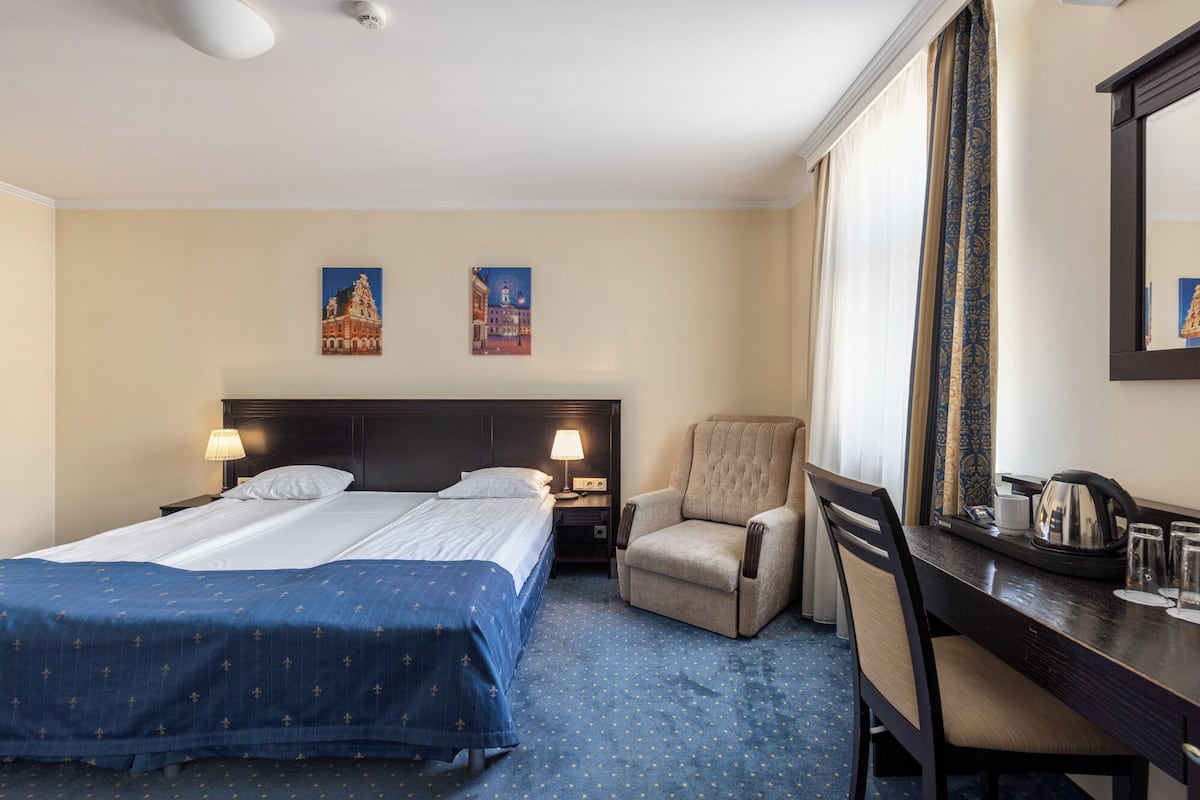 Habitación de hotel con cama, dos mesitas de noche con lámparas, una silla, escritorio con espejo y dos cuadros en la pared.