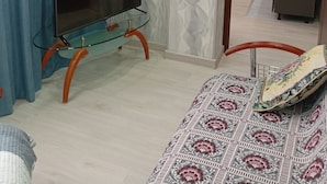 A bedroom with a glass TV stand, a bed with patterned bedding, and a view into a kitchen area.