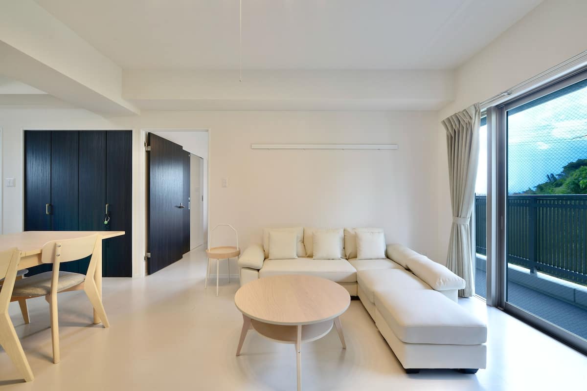 A modern living room with a sofa, a round table, and a dining table. There is a large window with curtains.