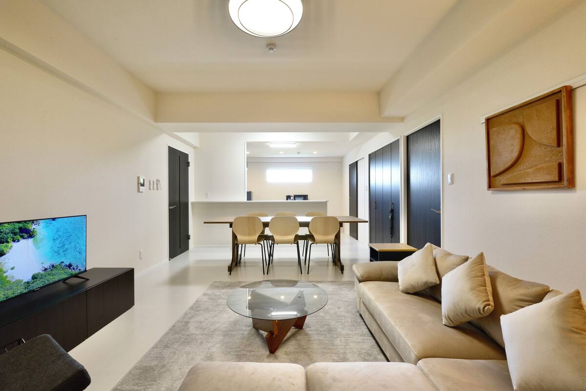 A modern living room with a flat-screen TV, a glass coffee table, a beige sofa, and a dining area with a table and chairs.