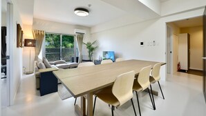 A modern living room with a large wooden dining table, beige chairs, a sofa, and a TV. - Riverside Chibana (Okinawa)