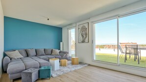 A modern living room with a grey sectional sofa, a small teal ottoman, and a wooden coffee table. A large window offers a view of a green lawn and buildings. - Stunning 1BR Flat with Terrace and private parking (Strasbourg)