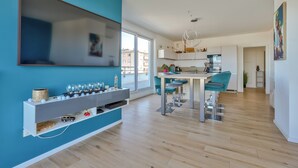 A modern kitchen with a central island, a dining area with teal chairs, and a large window providing a view of the city. - Stunning 1BR Flat with Terrace and private parking (Strasbourg)