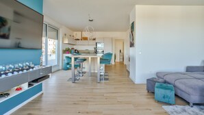 A modern living room with a grey sofa, a teal ottoman, and a kitchen area with a dining table and chairs. - Stunning 1BR Flat with Terrace and private parking (Strasbourg)