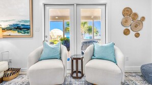 A room with two white armchairs, a hexagonal side table, a woven wall decoration, and a painting of a beach scene.