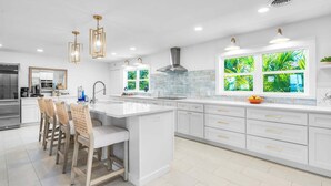 A modern kitchen with white cabinetry, a central island, pendant lighting, and a large window with a tropical view.