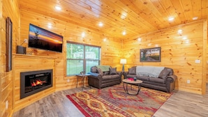 A cozy cabin interior with a fireplace, two sofas, a TV, and a large window.