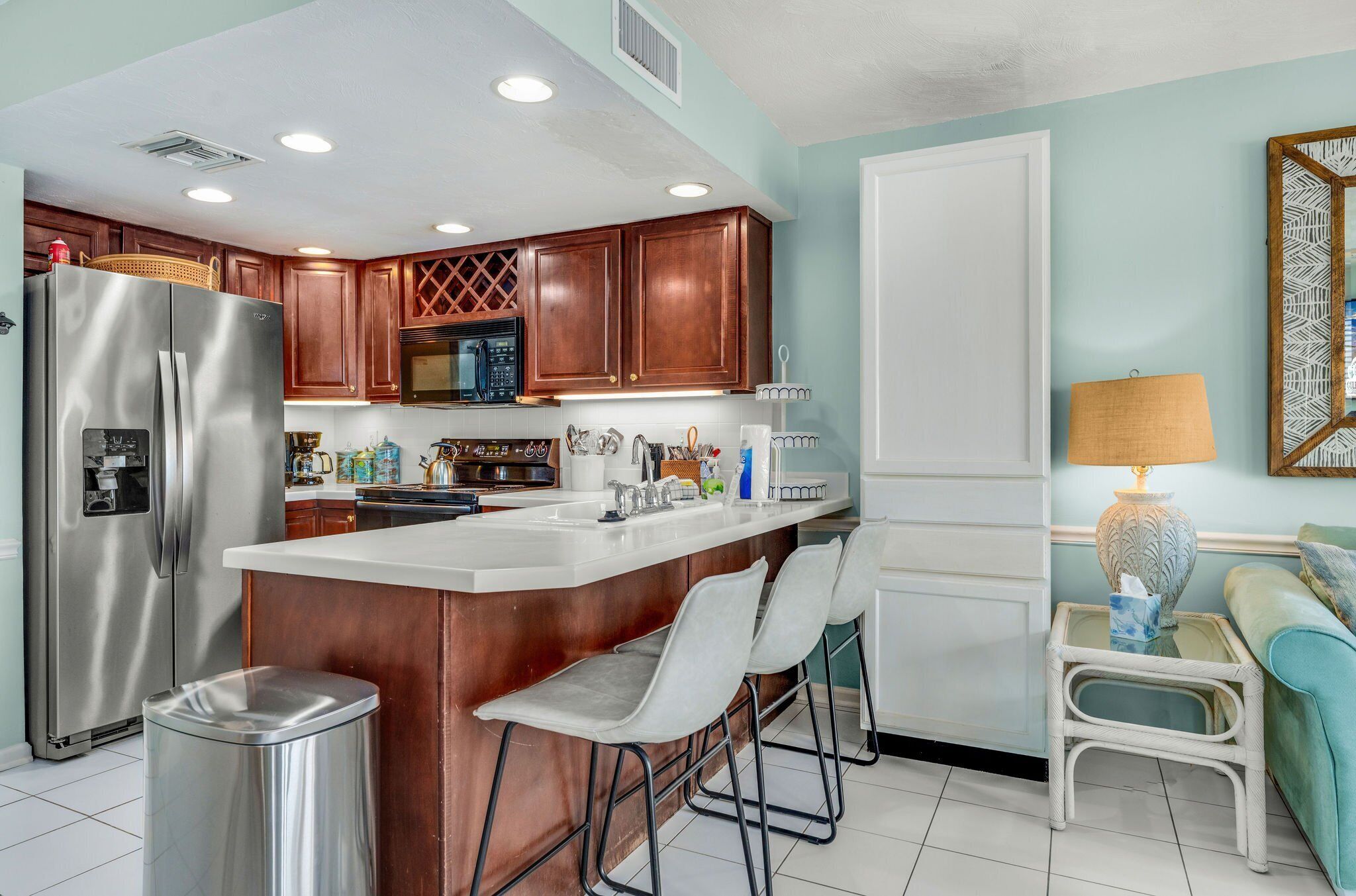 Una cocina moderna con refrigerador de acero inoxidable, armarios de madera, un espacio con taburetes de bar y una pequeña mesita con lámpara.
