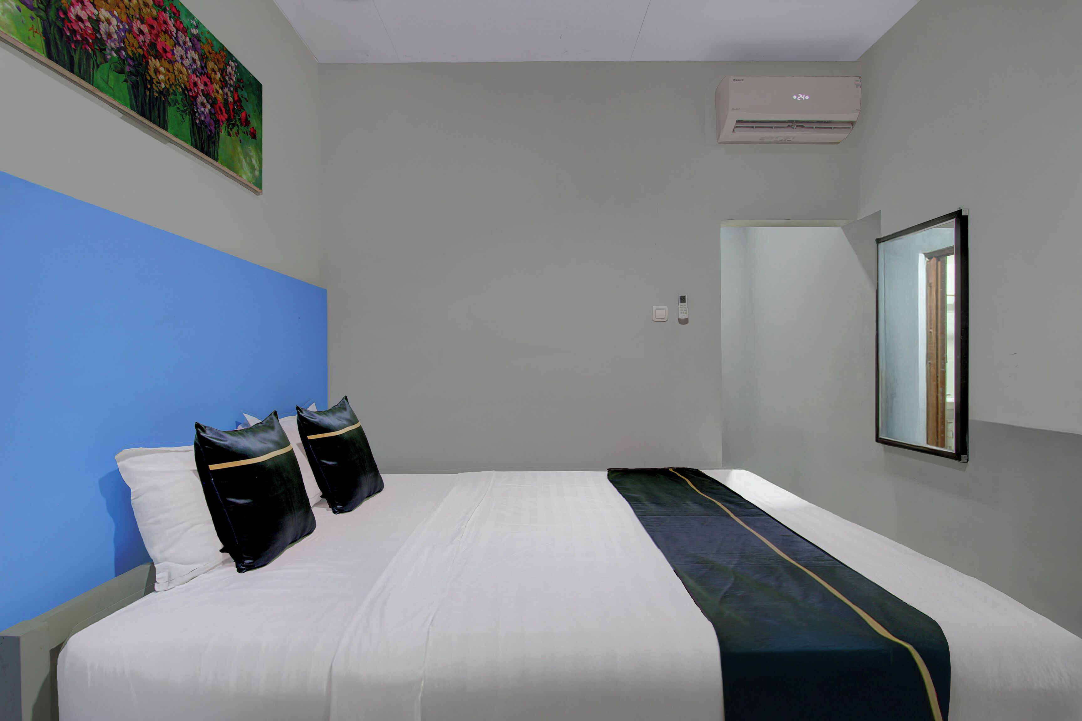 A hotel room with a bed, a blue accent wall, a painting, an air conditioning unit, and a mirror.