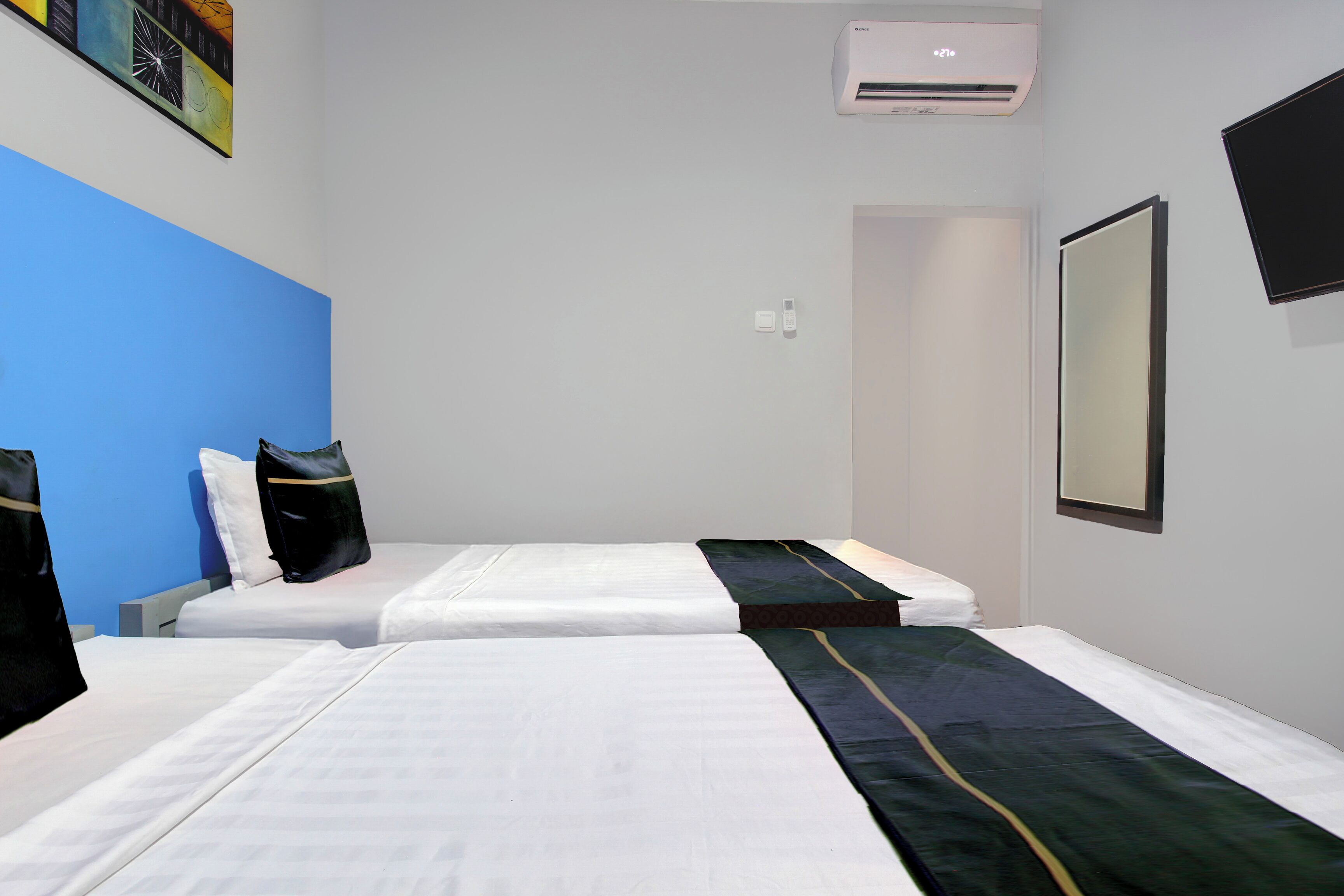 A hotel room with two beds, a blue accent wall, an air conditioning unit, a flat-screen TV, and a mirror.