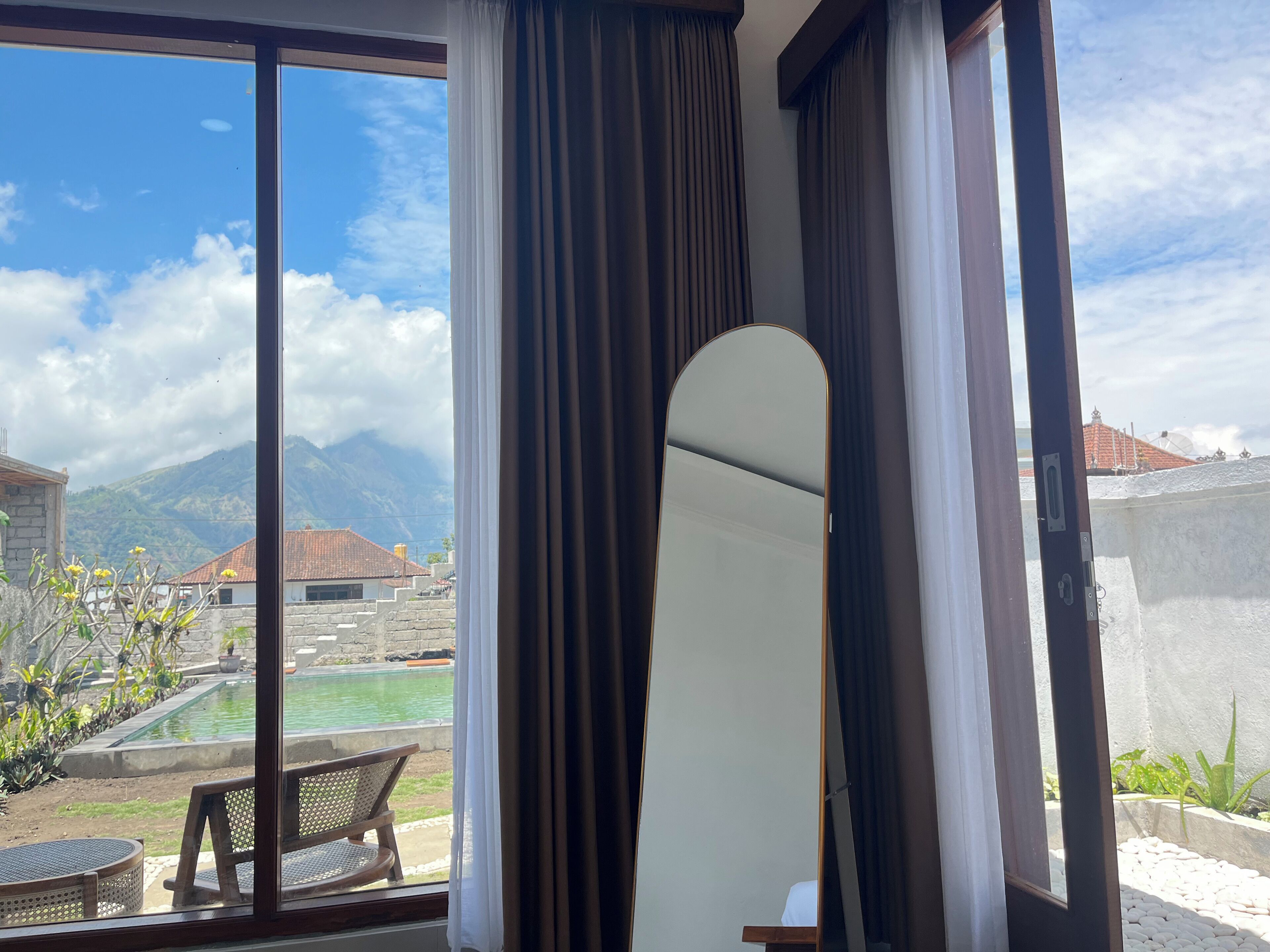 Ein Zimmer mit einer großen Schiebeglastür, die zu einem Poolbereich führt, einem Holzstuhl und Blick auf Berge sowie einen bewölkten Himmel.