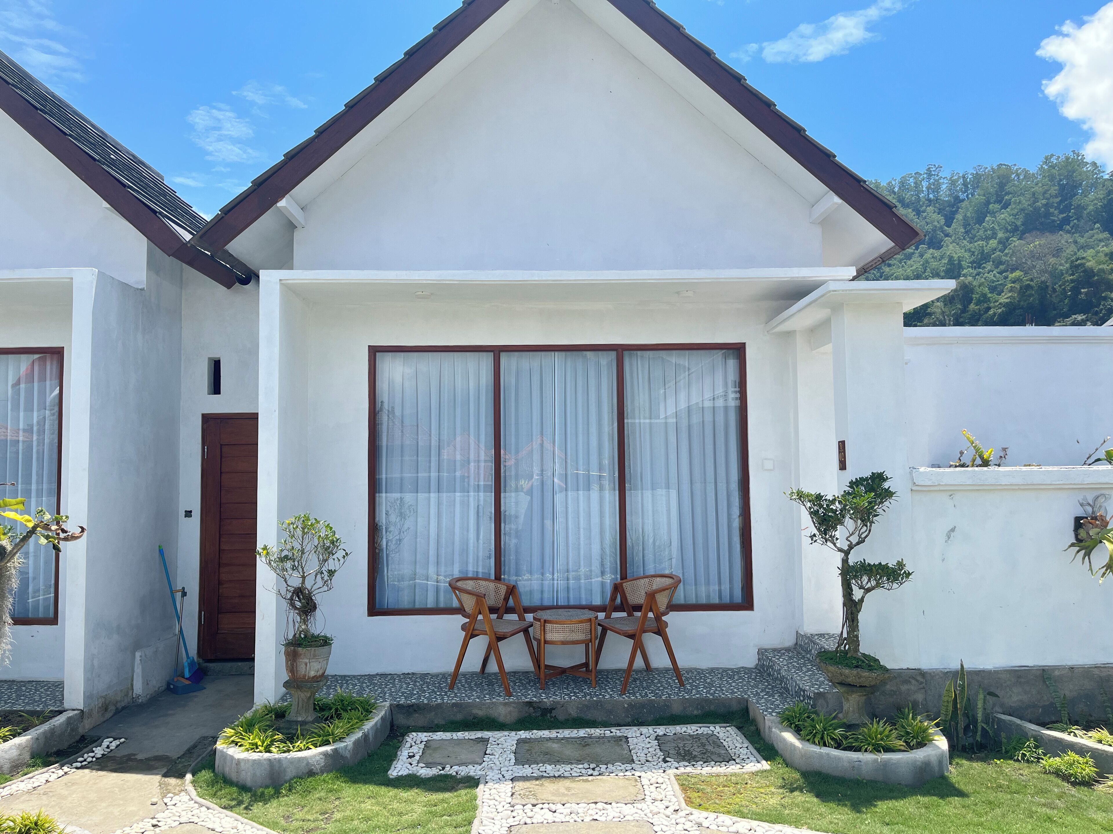 Una casa con fachada blanca, techo marrón y un ventanal grande con cortinas. Hay un pequeño jardín con un camino y plantas en macetas.