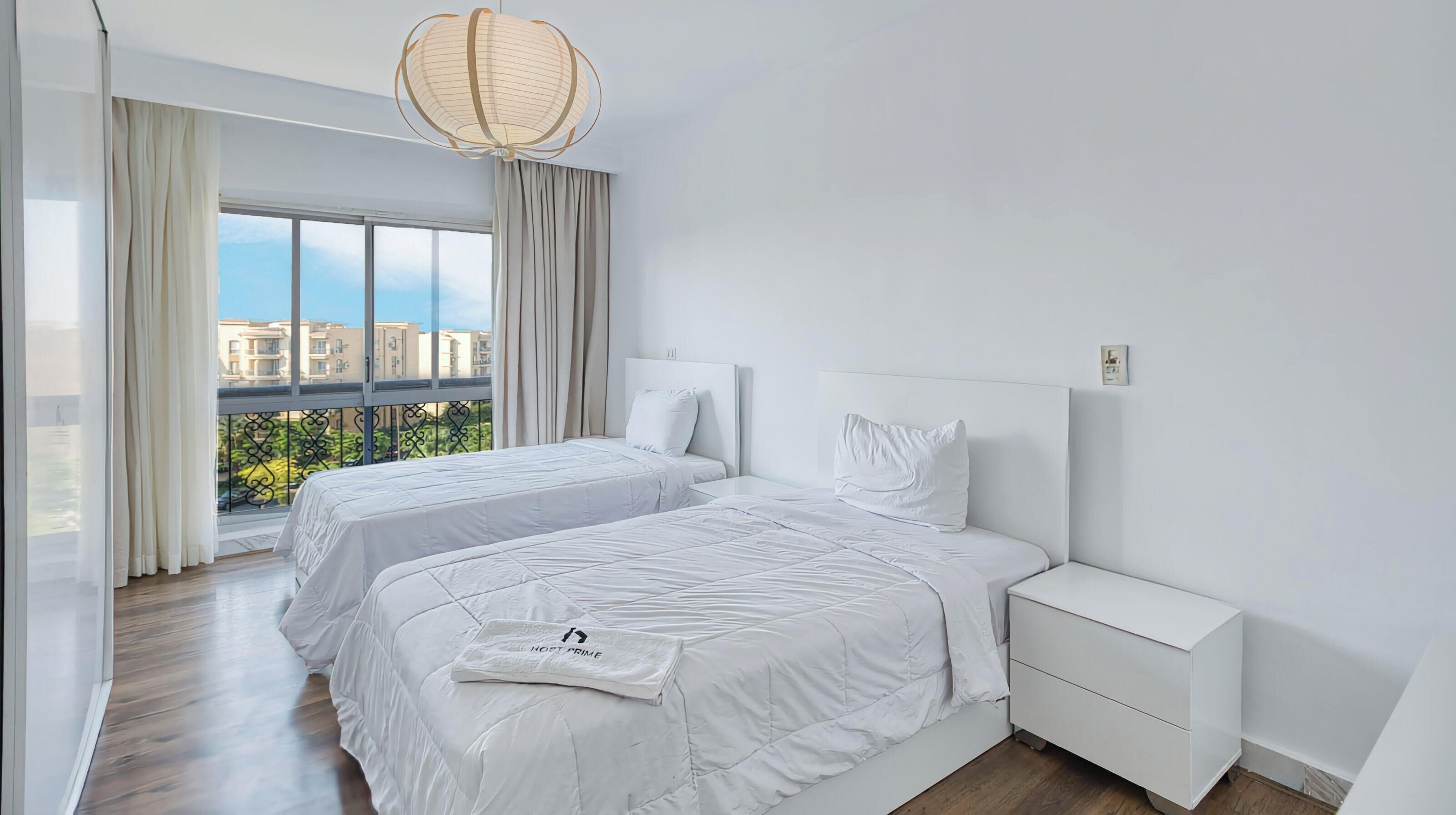 Una habitación de hotel moderna con dos camas, un ventanal con vistas, una mesita de noche blanca y una lámpara colgante.