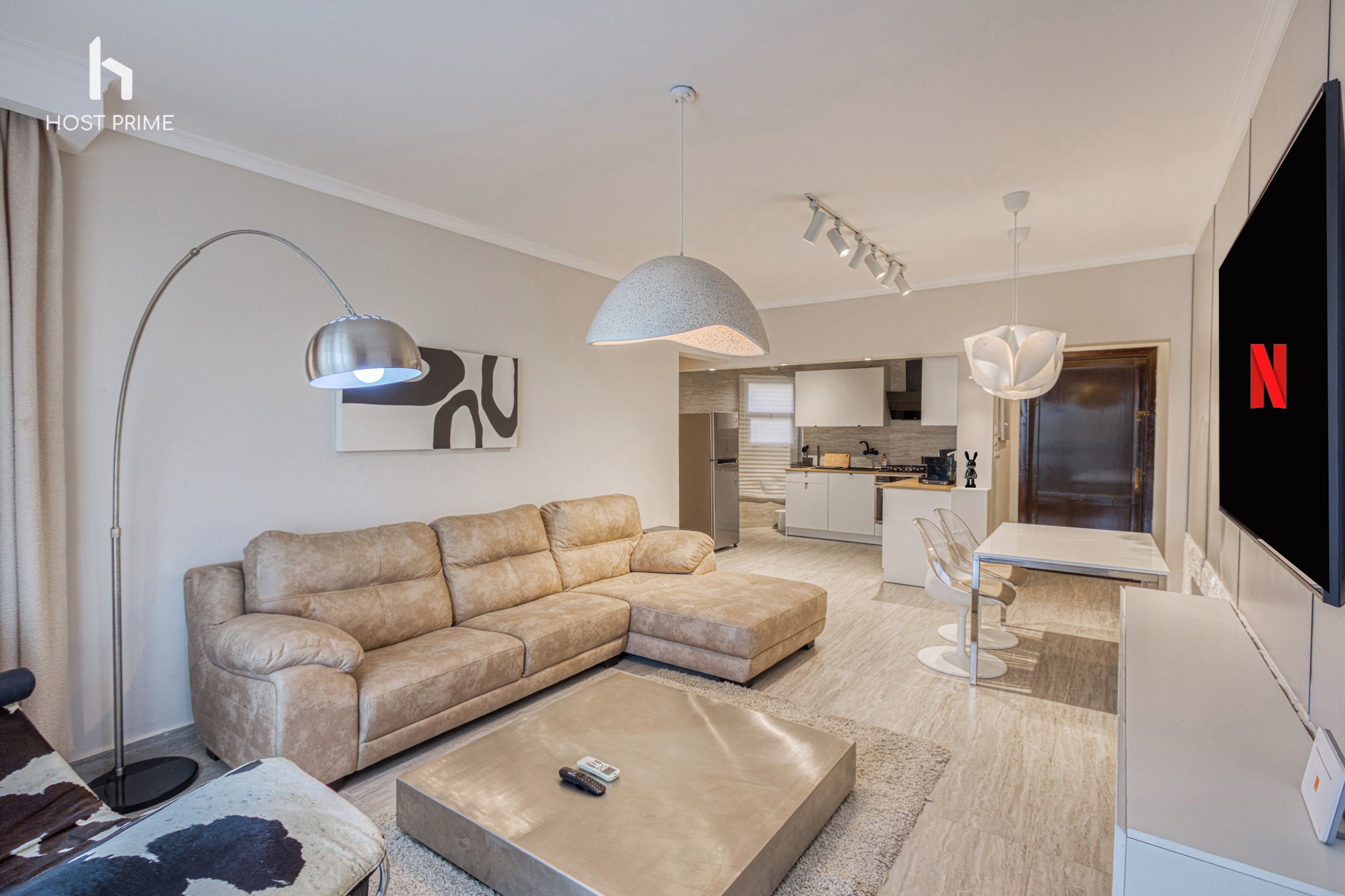 A modern living room with a sectional sofa, a coffee table, and a TV. There is a kitchen in the background with a dining area.