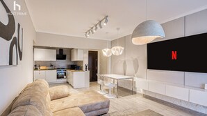 Uma sala de estar moderna com sofá, banqueta e área de jantar com mesa e cadeiras. É possível ver uma cozinha ao fundo, uma televisão na parede e um lustre no teto.