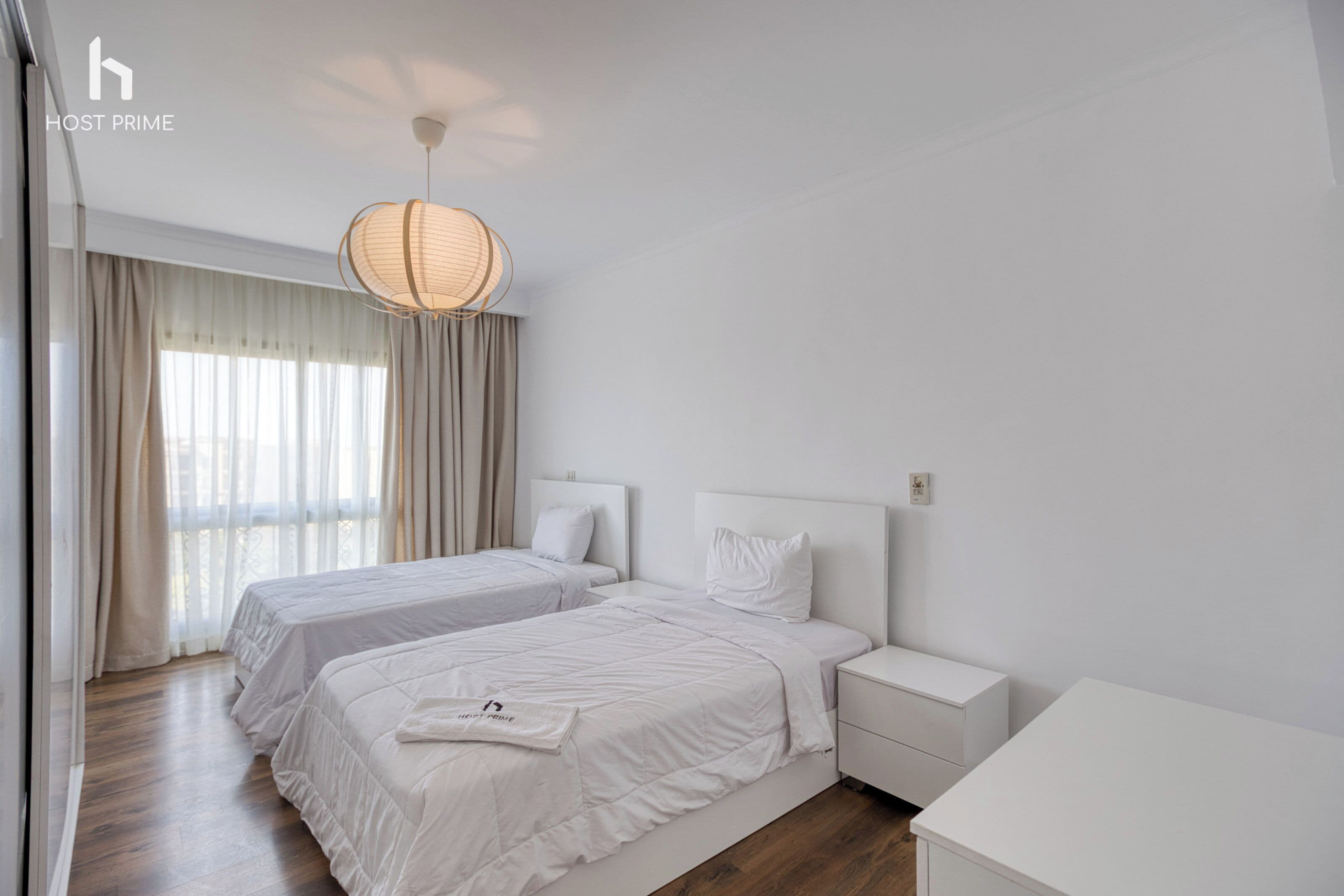A hotel room with two beds, a bedside table, a nightstand, and a large window with curtains.